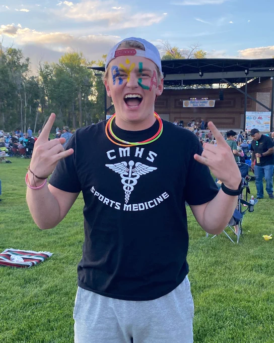 Person with face paint smiles broadly and makes a peace sign with both hands, wearing a RYLA t-shirt at an outdoor event.