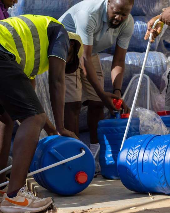 Three individuals filling blue barrels with water from a large container using a hose, likely involved in a humanitarian aid or disaster relief operation.