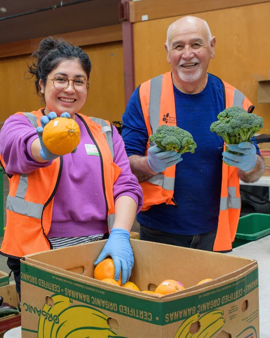 Two volunteers at a food bank, wearing safety vests and gloves, happily display fresh produce to the camera.
