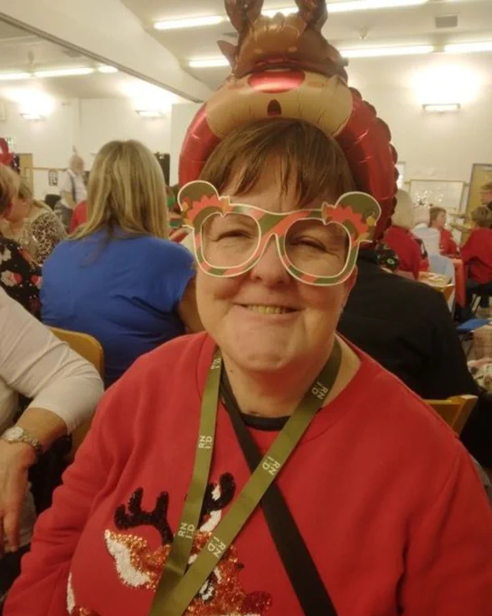 Woman wearing a red sweater with a reindeer design and a festive reindeer headband at a crowded indoor gathering.