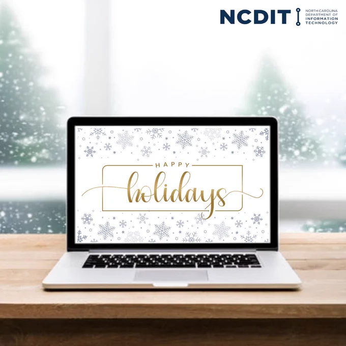 Laptop on a table displaying a "Happy Holidays" message with snowflake decorations on the screen, set against a backdrop of a window showing falling snow. 