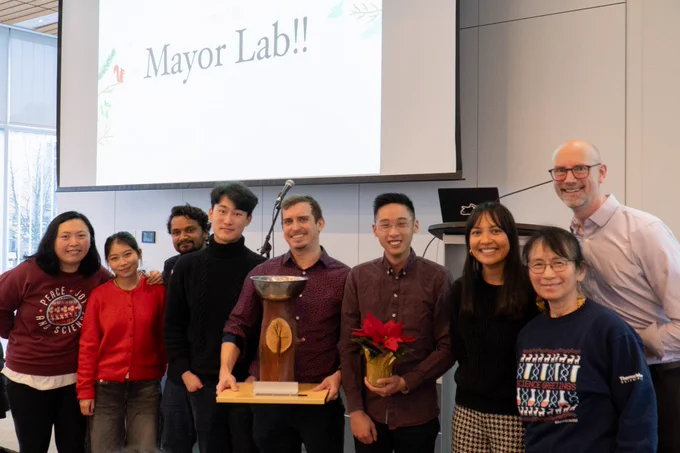 Members of the Mayor lab stand together, smiling while holding the trophy for winning the holiday video contest.