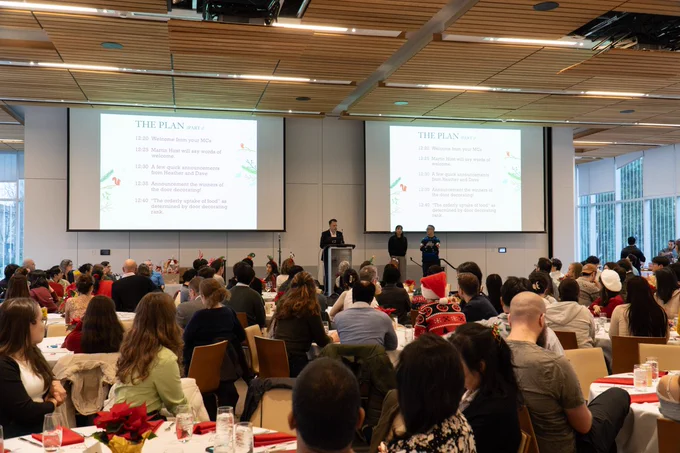 Dr. Martin Hirst stands at a podium on a small stage at the front of the room, delivering remarks to the room full of MSL members gathered around tables.