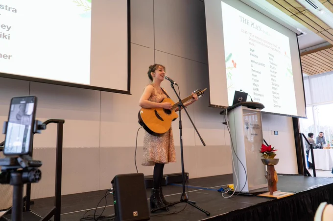 Postdoctoral fellow Dr. Marissa Lithopoulos stands at a microphone, guitar in hand, performing an original song for the attendees.