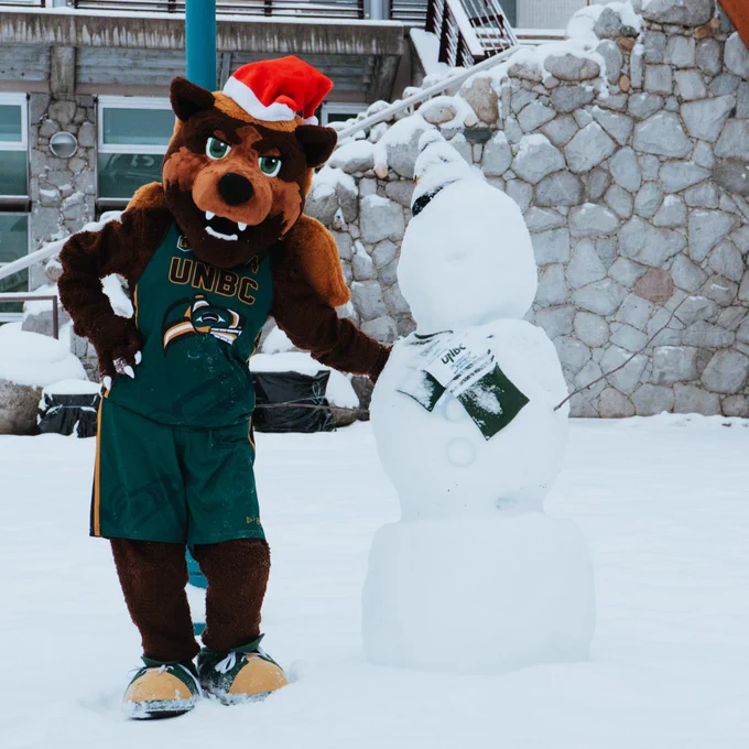 The UNBC Timberwolves mascot stands beside a snowman dressed in a UNBC t-shirt, in a snowy setting in front of a stone wall.
