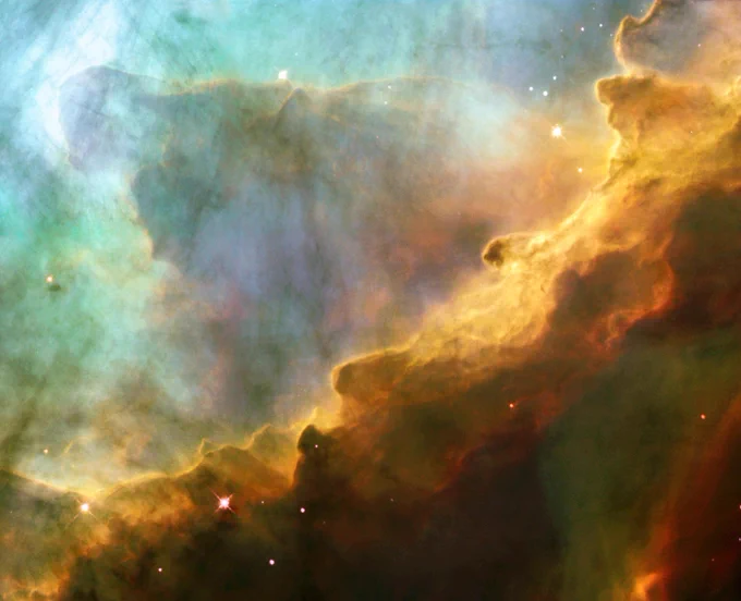 A colorful, painterly image of the gas and stars of Messier 17. The scene is split in two diagonally across the middle by different types of dust—a bright turquoise on the top-left half, and billows of ochre cascading down the bottom right half. Red stars are speckled about the scene. Credit: ESA, NASA, and J. Hester (Arizona State University)