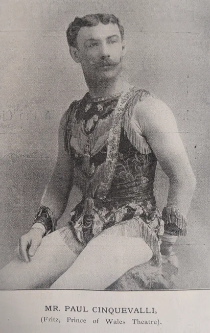 A sitting male figure with his head pointing to the right. His hair is parted and he has a waxed moustache. He is wearing a brocaded corset with an intricate  design. A medallion is around his neck falling down to his chest. His arms are bare and he has stockings on his legs. Text at the bottom of the photo indicates who the figure is and where they will be performing in Birmingham. 
