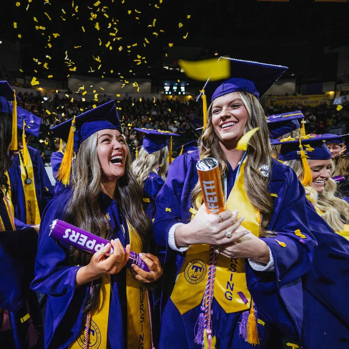 Graduates in purple caps and gowns hold confetti poppers as purple and gold confetti falls around them.