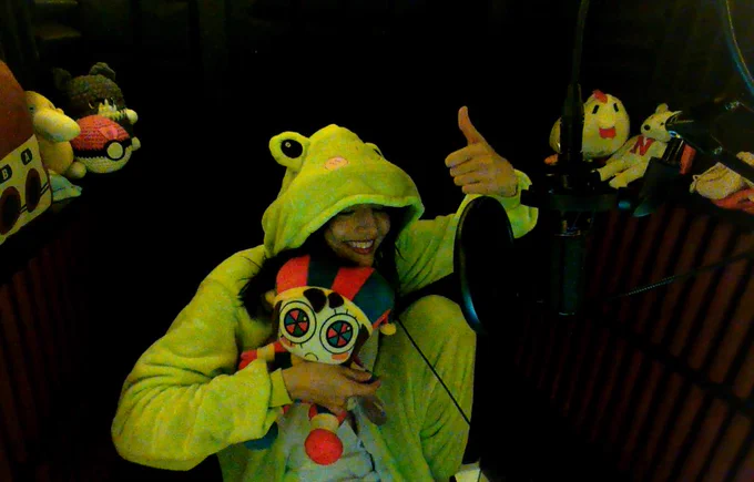 Skye Redden (some silly guy idk) in a green frog onesie, holding a Pomni plush, inside a recording booth, smiling with and giving a thumbs-up