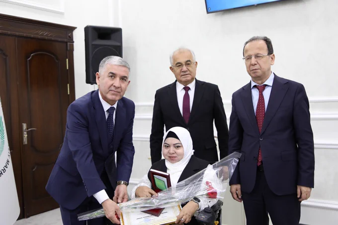 Our Chairperson Mukhabbat Rakhimova a woman in a wheelchair receives a certificate and flowers from officials during an award ceremony in a formal hall.