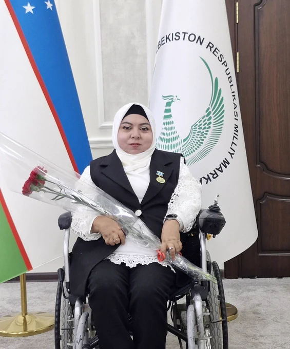A woman in a wheelchair Mukhabbat Rakhimova holding flowers and wearing a medal, seated in front of national and institutional flags.