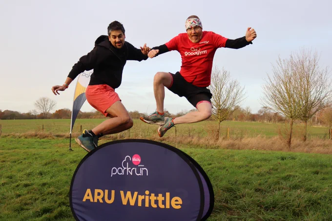 Two parkrunners, in black and red respectively,  joyfully jumping above a purple parkrun panel in a grassy field at ARU Writtle. 