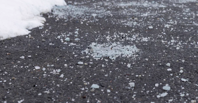 Close-up of icy salt granules scattered on a dark asphalt road beside melting snow.