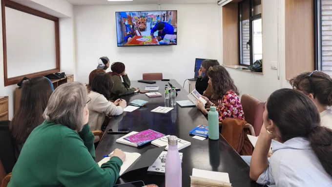 Sala de reuniones con varias personas sentadas alrededor de una mesa rectangular, observando una pantalla grande en l aparede donde se proyecta una imagen de una persona pintando en un taller de arte. Hay cuadernos, libros, botellas de agua y algunos teléfonos móviles sobre la mesa. La atmosfera es relajada y de concentración, pues están prestando atención a las explicaciones de las profesionales que trabajan en el proyecto Andar de Nones. La sala tiene grandes ventanas a la derecha y luz natural entrando.