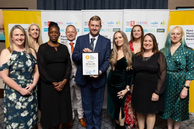 A group of nine happy and smiling people are in a line in front of the Kent Mental Wellbeing Awards banners. One in the middle is holding up the award winner's certificate.

