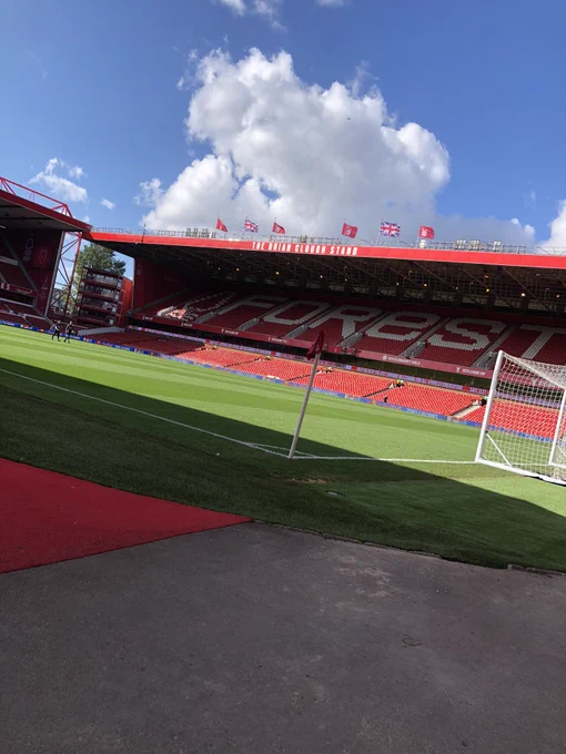 Photo taken inside Nottingham Forest’s City Ground Stadium.  The shot is taken on a bright summer day whilst the ground is empty, looking diagonally left onto the Brian Clough stand in the background. A red carpet type covering can be seen in the bottom left of the image with concrete to the right, Beyond is this is a red corner flag on top of a white pole, the lower part of the image is covered by shade. There is a full size goal and net to the right of the corner flag  and an immaculate, green pitch in the centre of the image. The empty Brian Clough Stand is awash with red seats apart from the word “Forest” which is depicted in white in the upper tier of the stand. At the very top of the image, above the Clough Stand, a huge fluffy white cloud hovers centrally amidst clear blue skies.