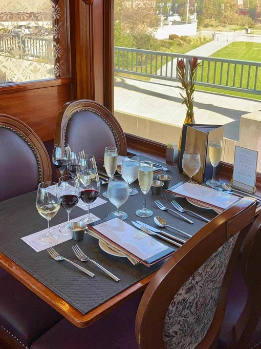 A dining table set for four, elegantly arranged with wine glasses, water glasses, and silverware, by a window overlooking a park.