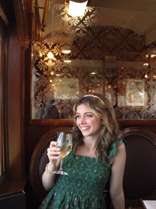 Person smiling, holding a glass of wine, seated inside a vintage-style dining car with ornate wallpaper.