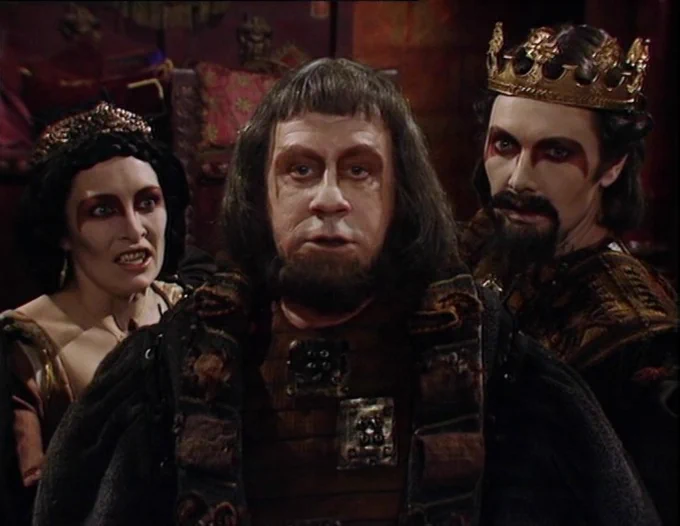 The Three Who Rule: Camilla, Aukon and Zargo, regal figures in dark red medieval garb standing in a richly decorated red room. Aukon stares ahead, his jaw clenched. Camilla and Zargo stand behind him, baring their teeth.
