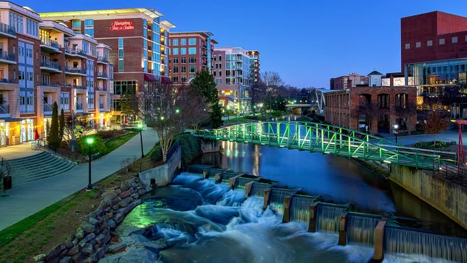 Evening view of Falls Park on the Reedy in downtown Greenville, South Carolina. The distinctive green Liberty Bridge arches over cascading waterfalls on the river, with modern buildings and warm lights reflecting in the water under a deep blue twilight sky.