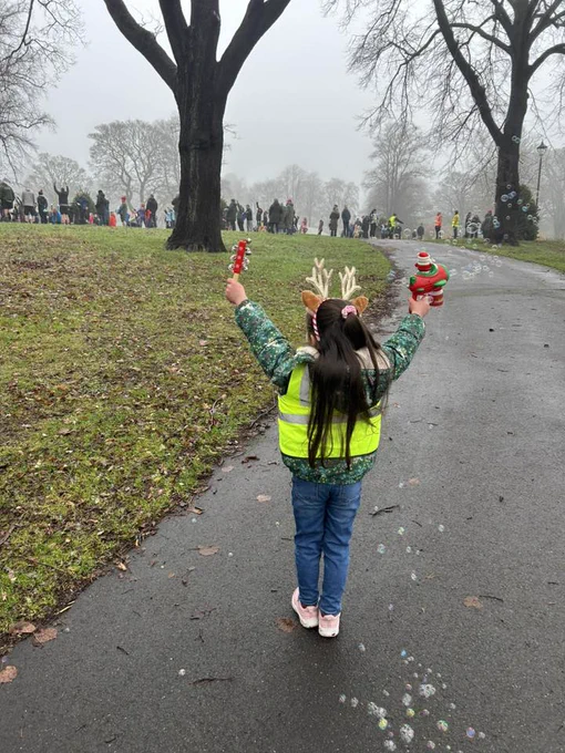 A junior parkrun volunteer, wearing a reindeer headband and holding two festive toys, cheering on parkrunners. 