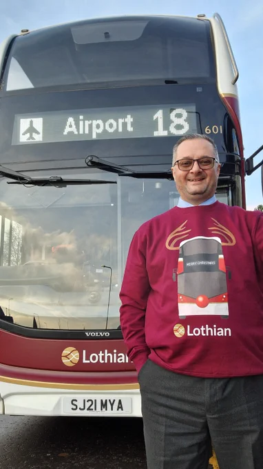 Person wearing a red Christmas Lothian sweatshirt stands in front of a double-decker bus displaying route 18 to the airport.
