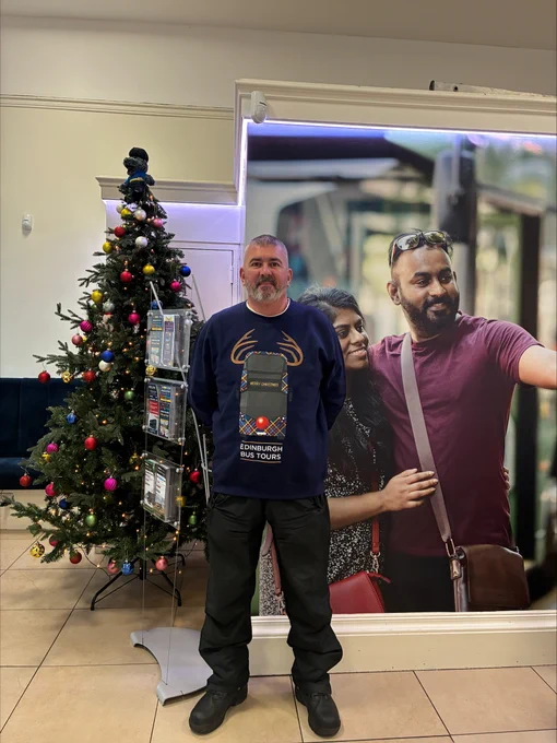 Man standing indoors near a decorated Christmas tree with colorful ornaments and lights.