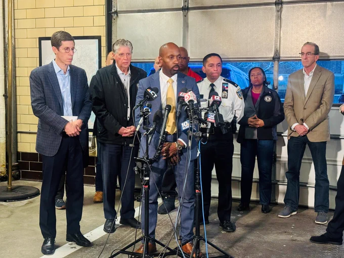 Group of officials and police officers standing behind a man speaking at a microphone during a press briefing in a garage-like setting.