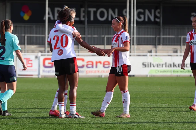 Atlanta Primus celebrates scoring against Royston Town with teammates, wearing the red and white home kit