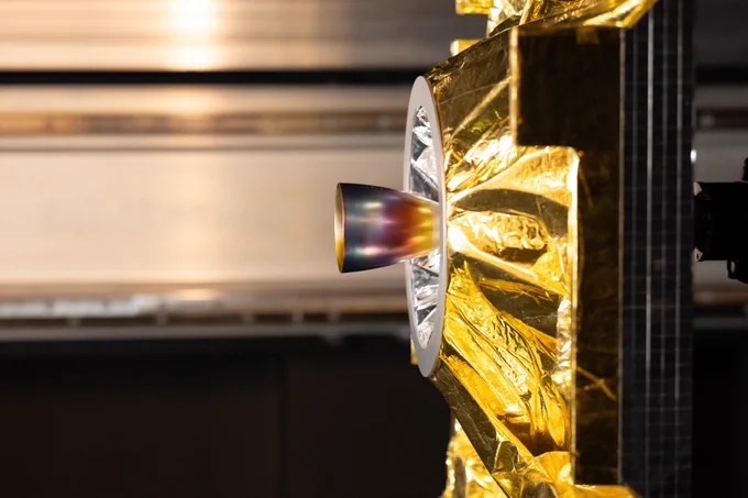 A sunlight lamp illuminates a satellite model covered in golden thermal insulation. A cup-shaped thruster extends from the model’s centre, reflecting a rainbow of colours. A few metres to the left, not captured in this image, a camera is brought closer and closer to the model, scanning the scene in this simulation of a rendezvous in space.