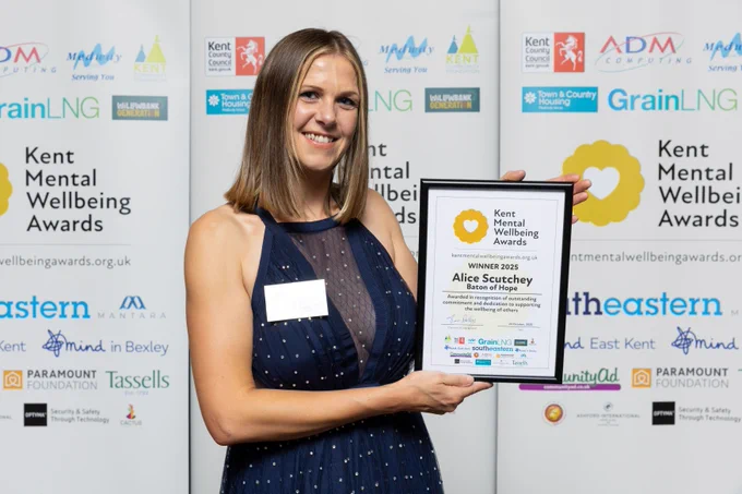 Alice is stood smiling in front of the Kent Mental Wellbeing Awards banner. She is holding the award winner's certificate.