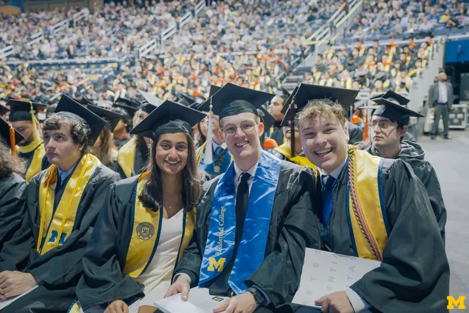Three graduates smiling while seated at 2025 Winter Commencement at Crisler Center.