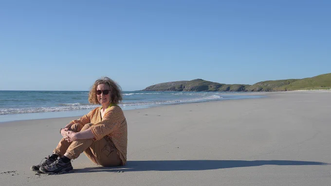 Photo of Professor Heather Lynch on a sandy Scottish beach with gentle waves and green hills in the background under a clear blue sky.