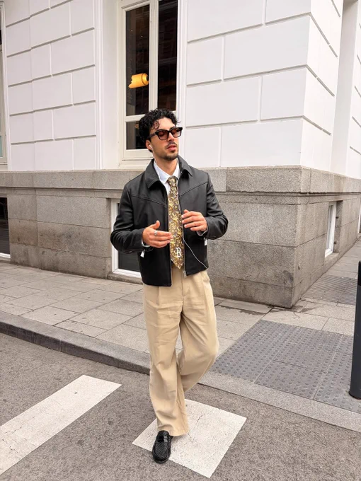 Street-style look featuring a black leather jacket, tailored beige trousers, and sunglasses, captured mid-stride in an urban setting.