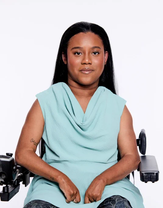 A woman with medium-dark skin and long straight black hair sits in a black wheelchair, facing the camera. She wears a sleeveless teal top with a cowl neckline and dark patterned pants; her hands rest in her lap, and a small tattoo is visible on her right forearm. The background is plain white.