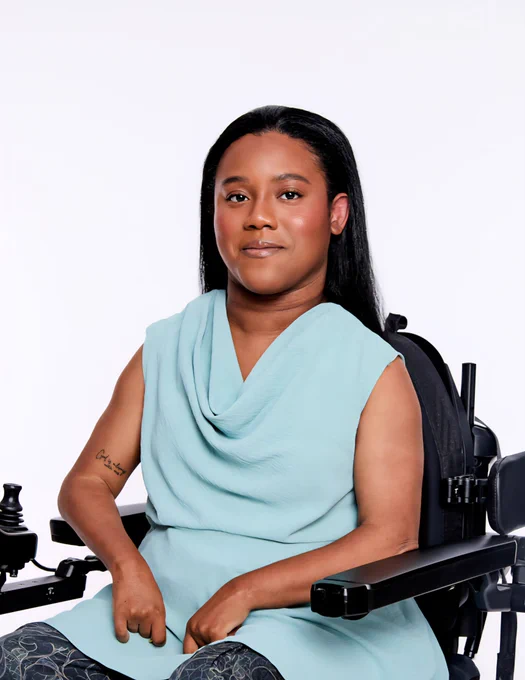 A woman with medium-dark skin and long straight black hair sits in a black wheelchair, facing the camera. She wears a sleeveless light blue top with a cowl neckline and dark patterned pants; her right hand rests on her lap, and a small black tattoo is visible on her right forearm. The background is plain white.