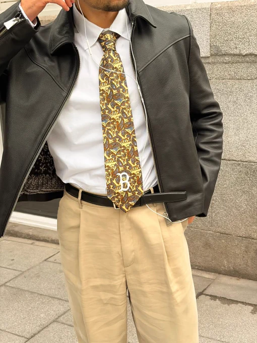 Detail shot of a street-style outfit featuring a black leather jacket, white dress shirt, paisley yellow tie, and tailored beige trousers.