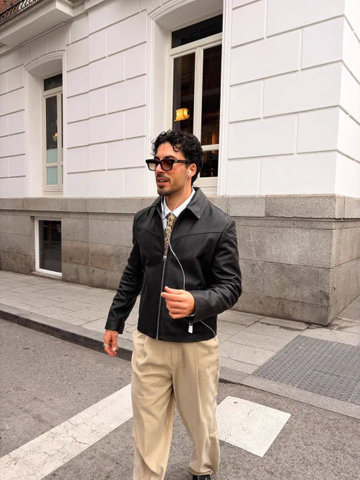 Street-style look featuring a black leather jacket, tailored beige trousers, and sunglasses, captured mid-stride in an urban setting.