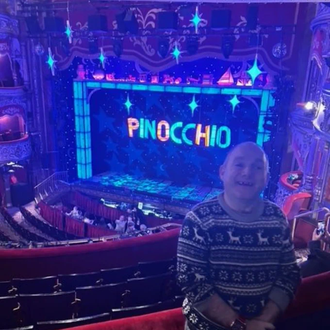 Man smiling with theatre stage in background, lit with colourful lights displaying 'PINOCCHIO' in large letters.