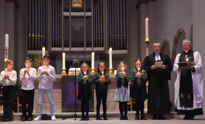 Photo credit: Hendrik zur Mühlen. School children are stood in a row in front of a Church Altar each holding an orange decorated with a red ribbon, a candle and sweets.  Two chaplains are stood with the children at the end of the row.