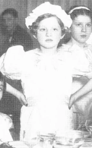 A vintage black and white photo of a girl, looking directly at the camera. Another girl is partially visible in the background.