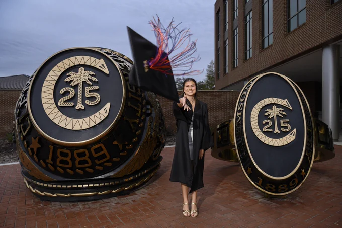 A person in academic regalia stands between two large Clemson Alumni Class Rings, smiling and mid-throwing a cap with a tassel in the air.