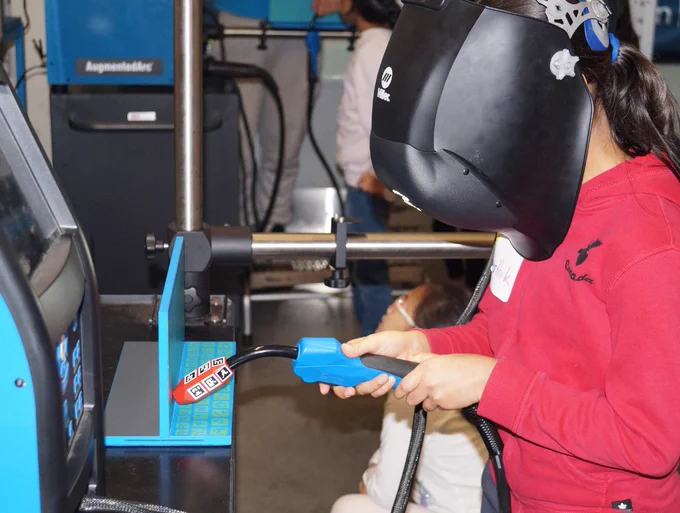 Youth wearing a welding helmet operates a virtual reality welding instrument, used for training