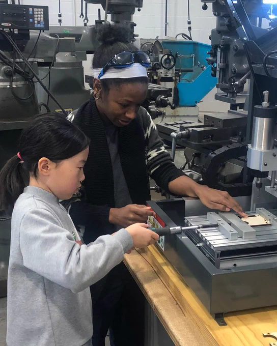 A STEM Role Model guides a youth operating equipment in a skilled trades workshop.