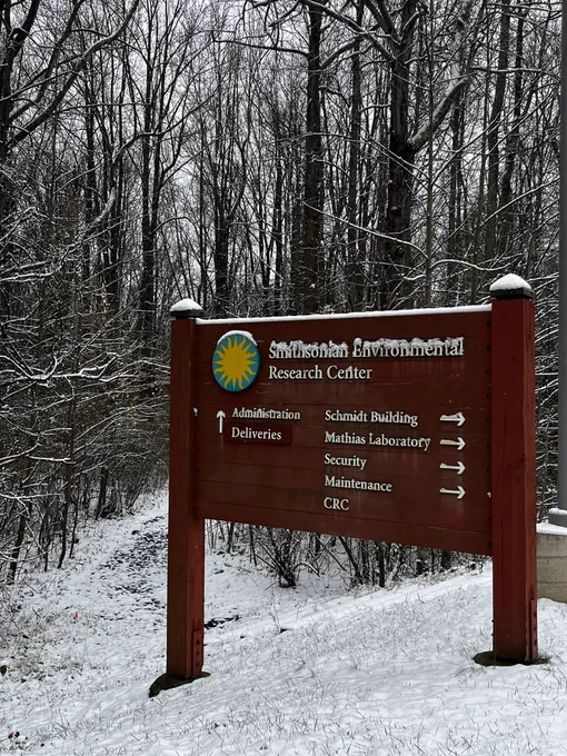 A wooden trailhead sign with the Smithsonian Environmental Research Center logo stands in front of a snow-covered forest. The top of the sign and its letters are lightly dusted in snow.