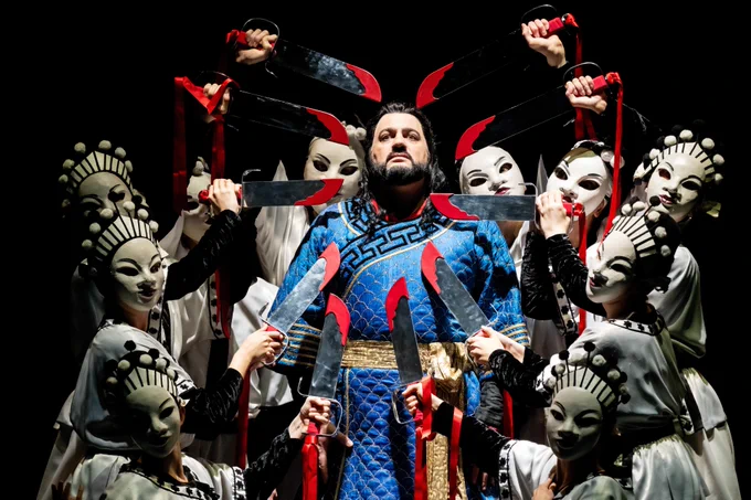 A person in a blue and red costume stands center stage surrounded by performers in white masks and black robes, holding red ribboned sticks, in a theatrical production.