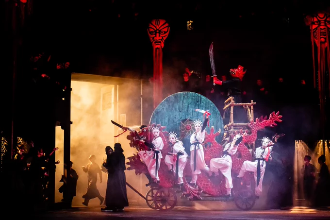 A theatrical performance featuring multiple performers in elaborate costumes around a decorative red and gold carriage, set against a backdrop with traditional motifs and dramatic lighting.