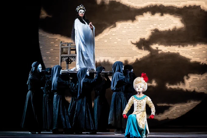 A theatrical stage scene from the opera "Turandot" featuring a performer in a white gown and mask standing elevated on a platform, surrounded by figures in dark cloaks, with a seated character in colorful, traditional attire.