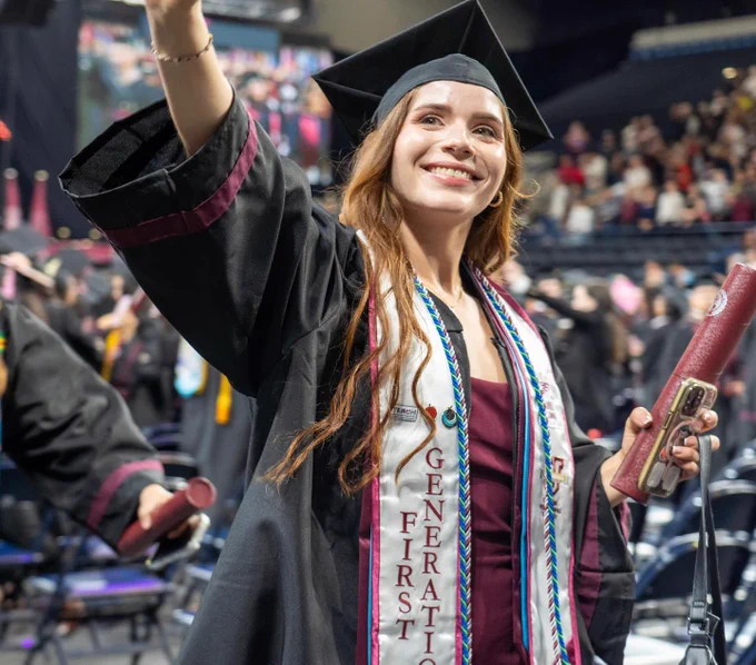 A smiling graduate wearing a black cap and gown waves to the crowd while holding a diploma during commencement. The graduate wears maroon academic regalia and honor cords, with fellow graduates and attendees visible in the background.