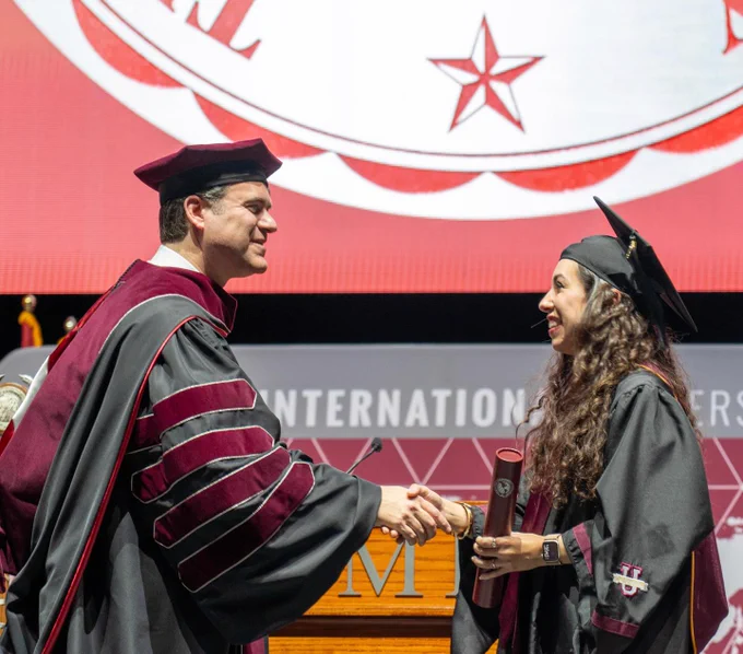 A graduate in cap and gown shakes hands with Texas A&M International University President Dr. Christopher Maynard on stage while holding a diploma. A large Texas A&M International University emblem is displayed on the screen behind them during the commencement ceremony.